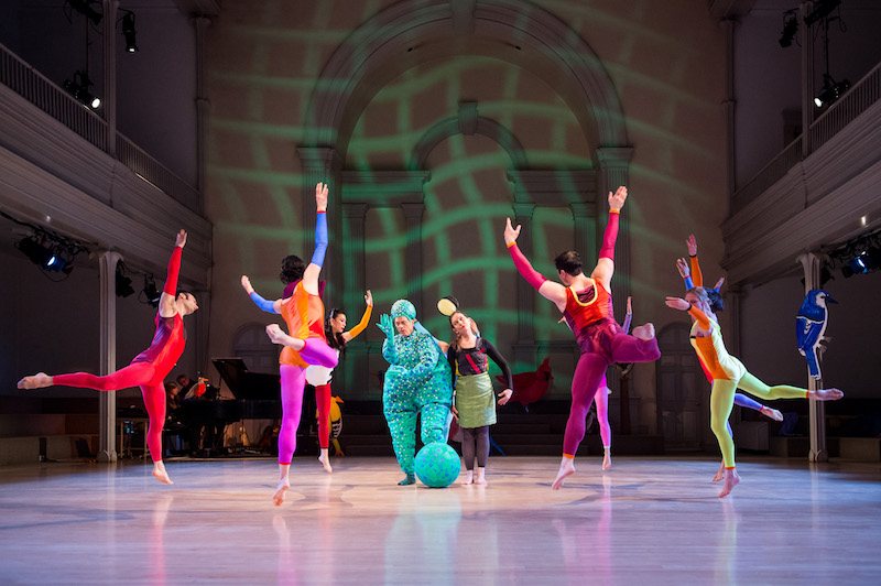 Dancers jump off one leg in a circle. Douglas Dunn in a puffy green costume resembling a monster. A woman in a green skirt and unitard stands next to him.